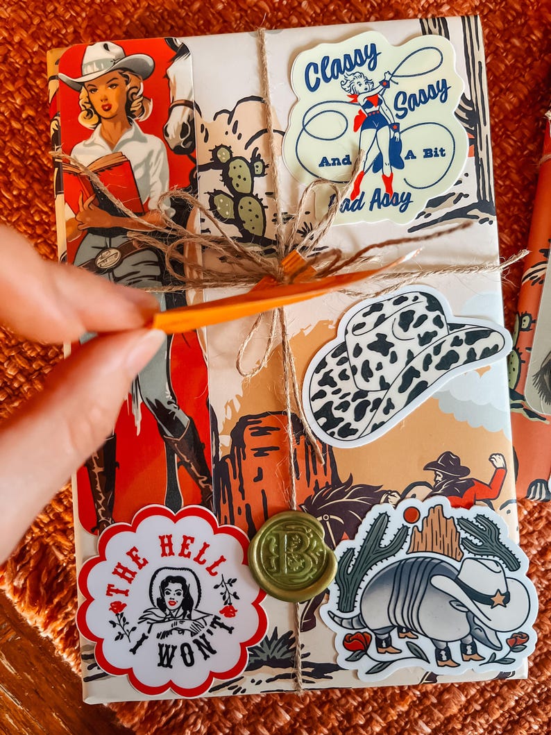 May include: A wrapped gift with a variety of colorful stickers. The stickers feature a cowgirl, a cactus, a armadillo, a hat, and a seal with the letter "B". The text on the stickers reads "Classy Sassy And A Bit Bad Ass", "The Hell I Won't", and "B".