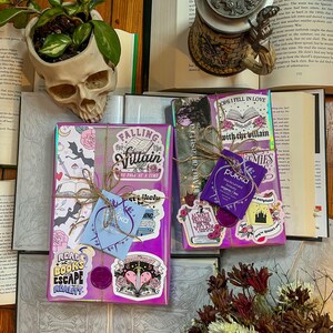 K&ouml;nnte beinhalten: Zwei verpackte B&uuml;cher mit bunten Aufklebern, eines mit dem Titel "Falling for the Villain" und das andere mit dem Titel "Oops I Fell in Love with the Villain". Die B&uuml;cher liegen auf einer Holzoberfl&auml;che mit einem Sch&auml;del-Pflanztopf und einem Buch mit einem Bierkrug.