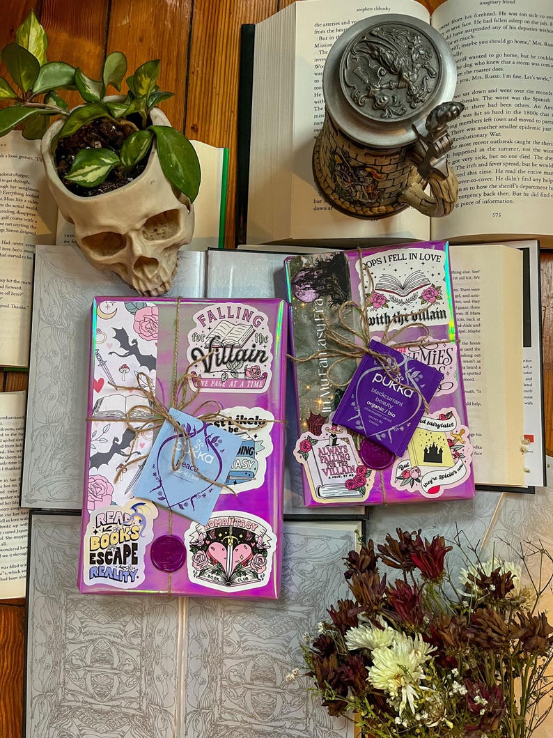 K&ouml;nnte beinhalten: Zwei verpackte B&uuml;cher mit bunten Aufklebern, eines mit dem Titel "Falling for the Villain" und das andere mit dem Titel "Oops I Fell in Love with the Villain". Die B&uuml;cher liegen auf einer Holzoberfl&auml;che mit einem Sch&auml;del-Pflanztopf und einem Buch mit einem Bierkrug.