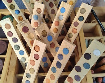 Può includere: Una collezione di tavolozze in legno, ognuna con una fila verticale di godet circolari di acquerelli in vari colori. Le tavolozze sono disposte in una scatola organizer in legno, alcune in posizione verticale e altre piatte, mostrando sfumature di blu, marrone, giallo e verde.