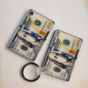 May include: Two keychains shaped like $100 bills with a silver ring attached. The bills have a blue and white design with a portrait of Benjamin Franklin.