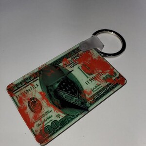 May include: A green and white $100 bill keychain with a red blood splatter design. The bill features a portrait of Benjamin Franklin with a bandana over his face.
