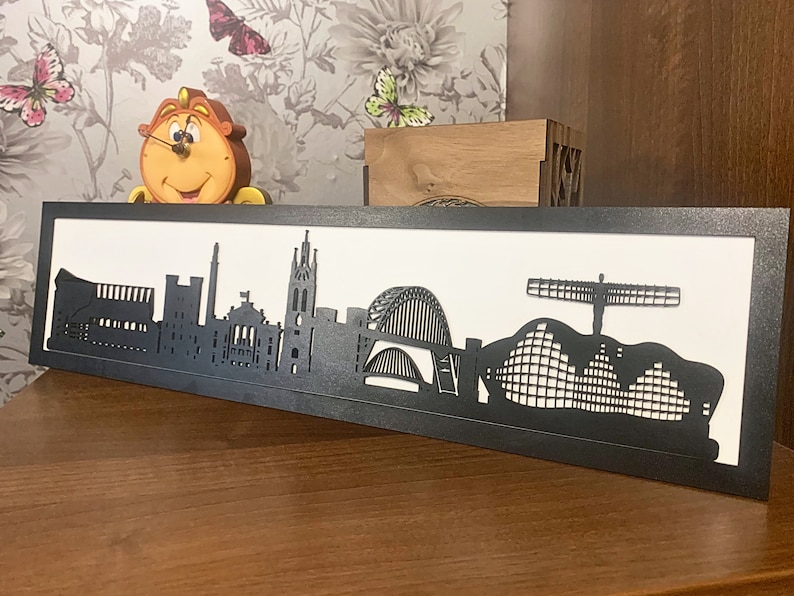 May include: Black and white silhouette of the Newcastle skyline, including the Tyne Bridge, the Sage Gateshead, and the Angel of the North. The silhouette is mounted in a black frame.