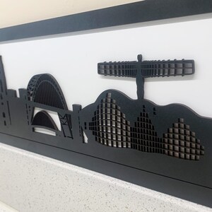 May include: Black and white silhouette artwork of the city skyline of Manchester, England. The artwork features iconic landmarks such as the Manchester Town Hall, the Arndale Centre, and the Angel of the North.