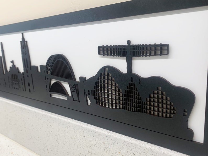 May include: Black and white silhouette artwork of the city skyline of Manchester, England. The artwork features iconic landmarks such as the Manchester Town Hall, the Arndale Centre, and the Angel of the North.