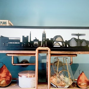 May include: Black silhouette of a city skyline framed in a black frame, sitting on a shelf with copper pipes. The shelf is part of a copper pipe cart with a glass terrarium and copper-colored pears on the lower shelf.