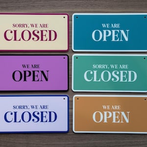 May include: Six rectangular signs with rounded corners and two small holes at the top. The signs feature the text "SORRY, WE ARE CLOSED" or "WE ARE OPEN" in various colour combinations including cream, teal, purple, green, light blue, and brown.