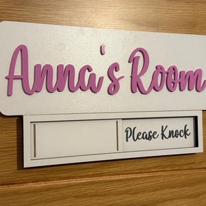 May include: A rectangular white door sign featuring the text Anna's Room in raised pink script lettering. Below the main sign is a sliding mechanism with a white panel displaying the black text Please Knock on the right side.