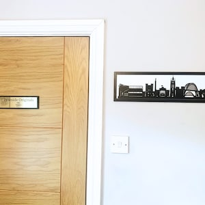 May include: A wooden door with a sign that reads "TyneSide Originals" and a black metal silhouette of a city skyline on the wall behind it.