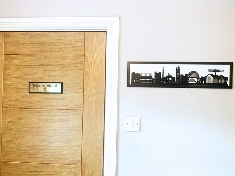 May include: A wooden door with a sign that reads "TyneSide Originals" and a black metal silhouette of a city skyline on the wall behind it.
