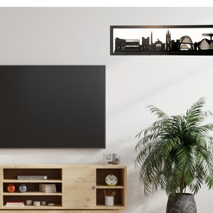 May include: Black metal silhouette of a city skyline, mounted on a white wall above a wooden entertainment center with a flat screen TV and a potted plant.