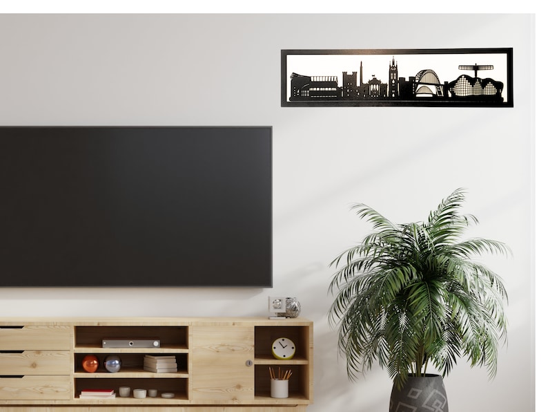 May include: Black metal silhouette of a city skyline, mounted on a white wall above a wooden entertainment center with a flat screen TV and a potted plant.