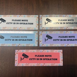 May include: Rectangular signs with the text "PLEASE NOTE CCTV IS IN OPERATION" and a security camera graphic. The signs have different colored backgrounds: white, silver, light blue, purple, and red. Each sign has a decorative border with a pattern of baked goods.