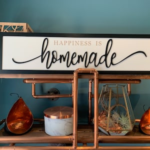 May include: A black framed sign with a white background and the text "Happiness is homemade" in black cursive lettering. The sign is on a shelf made of copper pipes.