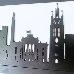 May include: Black silhouette of a city skyline with tall buildings and a church steeple. The skyline is made of wood and is mounted on a white background.