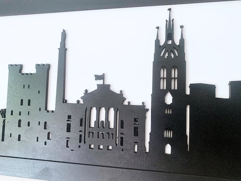 May include: Black silhouette of a city skyline with tall buildings and a church steeple. The skyline is made of wood and is mounted on a white background.