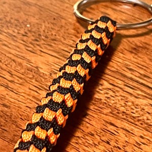 May include: A keychain featuring a cylindrical paracord braid in a repeating pattern of bright orange and black nylon cord. The braided section is attached to a circular silver-toned metal split ring at the top, creating a durable accessory.
