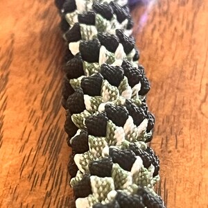 May include: A paracord lanyard featuring a complex, multi-layered weave pattern. The cord is constructed from interwoven strands in solid black, olive green, and white, creating a dense, textured, and cylindrical design with a repeating knot structure.
