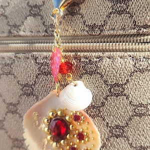 Peut inclure: Un porte-clés coquillage avec des détails dorés et un fermoir mousqueton. La coquille est ornée de pierres précieuses rouges et de perles dorées. Des breloques supplémentaires comprennent une feuille rose et des perles rouges. Fond marron à motifs.