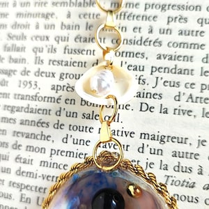 Peut inclure: Un collier à chaîne torsadée dorée avec un pendentif composé d'un petit coquillage blanc contenant une perle, relié par des anneaux dorés à un coquillage plus grand bordé de fil doré, orné d'une perle noire et de petites perles dorées.