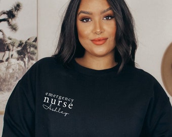May include: Black crew neck sweatshirt with the text "emergency nurse Ashley" in white script. The sweatshirt is a solid black colour and has long sleeves. The image also shows a hat and a tote bag.