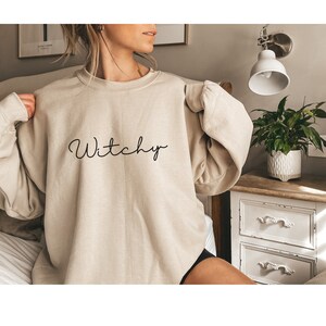 May include: A beige sweatshirt with the word "Witchy" written in black cursive script.