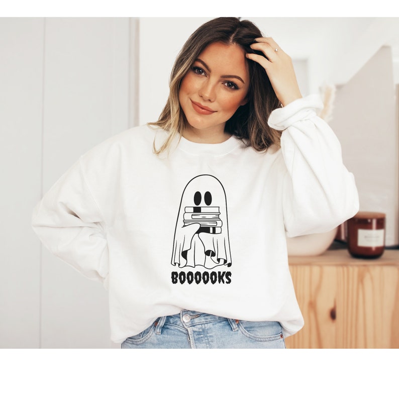 May include: White sweatshirt with a black and white illustration of a ghost holding a stack of books. The text "BOO-OO-OO-OOKS" is printed below the ghost.
