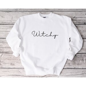 May include: White sweatshirt with the word "Witchy" written in black cursive script.