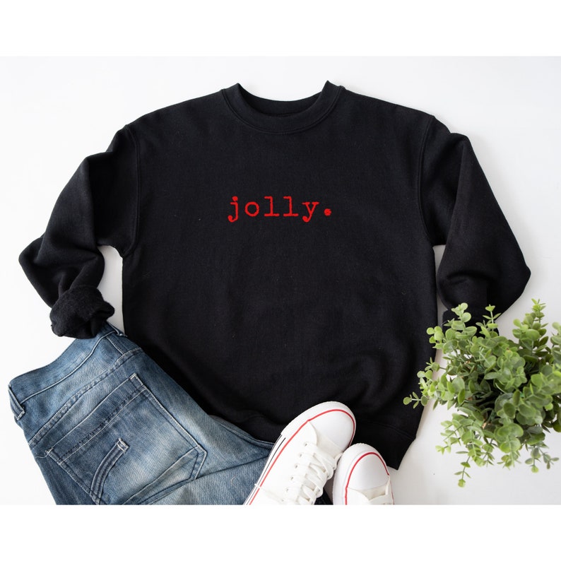 May include: Black sweatshirt with the word "jolly" in red embroidered on the front. The sweatshirt is laying on a white surface with a pair of white sneakers and blue jeans.
