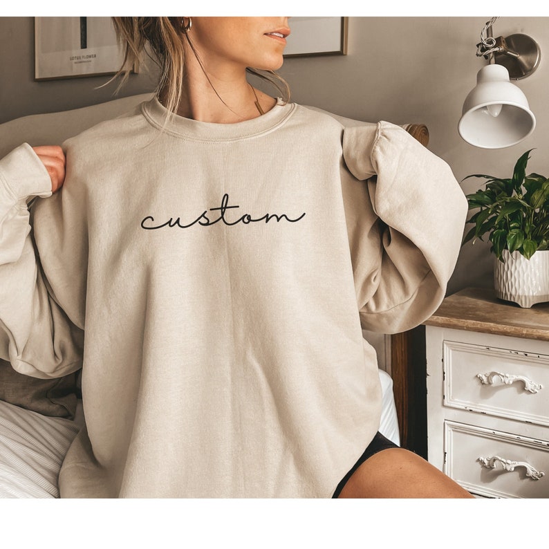 Puede incluir: Una sudadera beige de gran tama&ntilde;o con la palabra "custom" bordada en negro en la parte delantera.