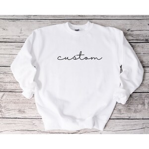 Puede incluir: Sudadera blanca de cuello redondo con la palabra "custom" escrita en cursiva negra.