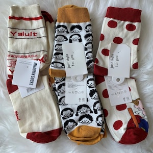 May include: Four pairs of white socks with different designs. One pair has red and white stripes with the text "Yalult" and Japanese characters. Another pair has a yellow and white design with a repeating pattern of cartoon faces. The third pair has red polka dots and the fourth pair has a red and white stripe with a cartoon character.