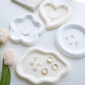 May include: Three white ceramic jewelry dishes in different shapes: a heart, a cloud, and a flower. The dishes are on a white surface and are decorated with black speckles. The dishes are filled with jewelry, including a pearl necklace, a pair of gold earrings, and a gold ring.