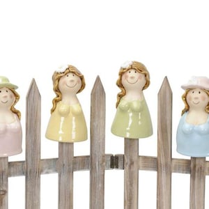 Op de afbeelding: Vier keramische beeldjes van vrouwen met verschillende gekleurde jurken en hoeden, die op een houten schutting staan.