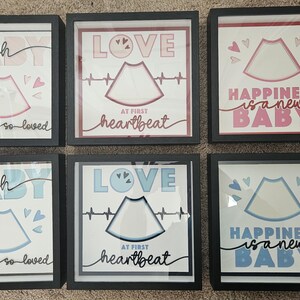 May include: Six framed art prints with baby-themed designs. Each print features a sonogram image, hearts, and text like "Oh Baby," "Love at first heartbeat," and "Happiness is a new baby." The frames are black.
