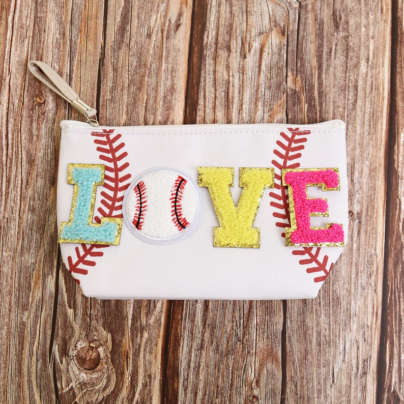 Puede incluir: Bolsa de lona blanca con tem&aacute;tica de b&eacute;isbol con la palabra "LOVE" en letras coloridas y texturizadas. La "L" es azul claro, la "V" es amarilla y la "E" es rosa. Un parche de b&eacute;isbol est&aacute; en el centro. La bolsa tiene una correa de mu&ntilde;eca y costuras rojas.