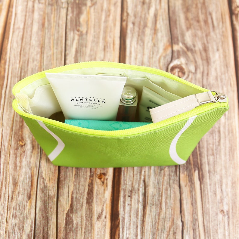 Puede incluir: Un neceser verde lima con un dise&ntilde;o de pelota de tenis blanca, abierto para revelar productos para el cuidado de la piel y maquillaje. La bolsa tiene una cremallera amarilla y una leng&uuml;eta blanca. La bolsa est&aacute; sobre una superficie de madera.