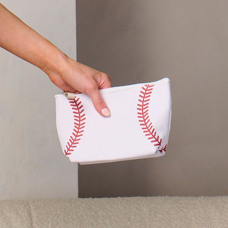 Puede incluir: Bolsa de cosm&eacute;ticos blanca con tem&aacute;tica de b&eacute;isbol con detalles de costura rojos. La bolsa tiene un cierre de cremallera dorado y est&aacute; dise&ntilde;ada para parecerse a una pelota de b&eacute;isbol. Un accesorio elegante para los fan&aacute;ticos del deporte.