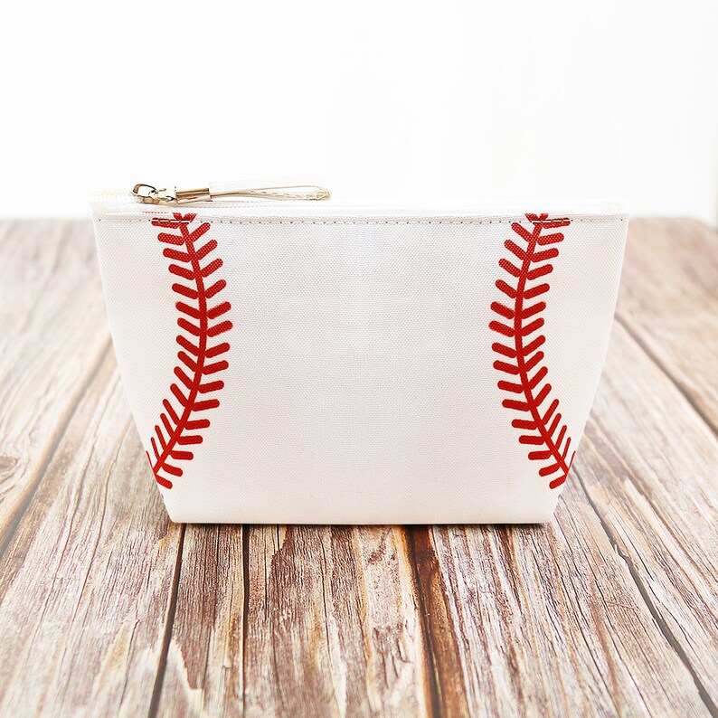 Puede incluir: Bolsa de cosm&eacute;ticos blanca con tem&aacute;tica de b&eacute;isbol con detalles de costuras rojas. La bolsa tiene un cierre de cremallera y una peque&ntilde;a correa de mu&ntilde;eca. El dise&ntilde;o imita el aspecto de una pelota de b&eacute;isbol.