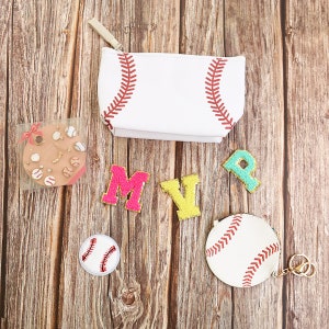 Puede incluir: Una colecci&oacute;n de accesorios con tem&aacute;tica de b&eacute;isbol sobre una superficie de madera. Incluye una bolsa blanca cosida con costuras de b&eacute;isbol, un monedero redondo con un llavero dorado, parches que deletrean "MVP" y un parche con forma de b&eacute;isbol. Tambi&eacute;n, un paquete de galletas con tem&aacute;tica de b&eacute;isbol.