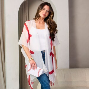 Puede incluir: Un kimono blanco con detalles rojos, combinado con una camiseta blanca y vaqueros azules. Se sostiene un clutch blanco con costuras rojas de b&eacute;isbol. El conjunto es informal y elegante.