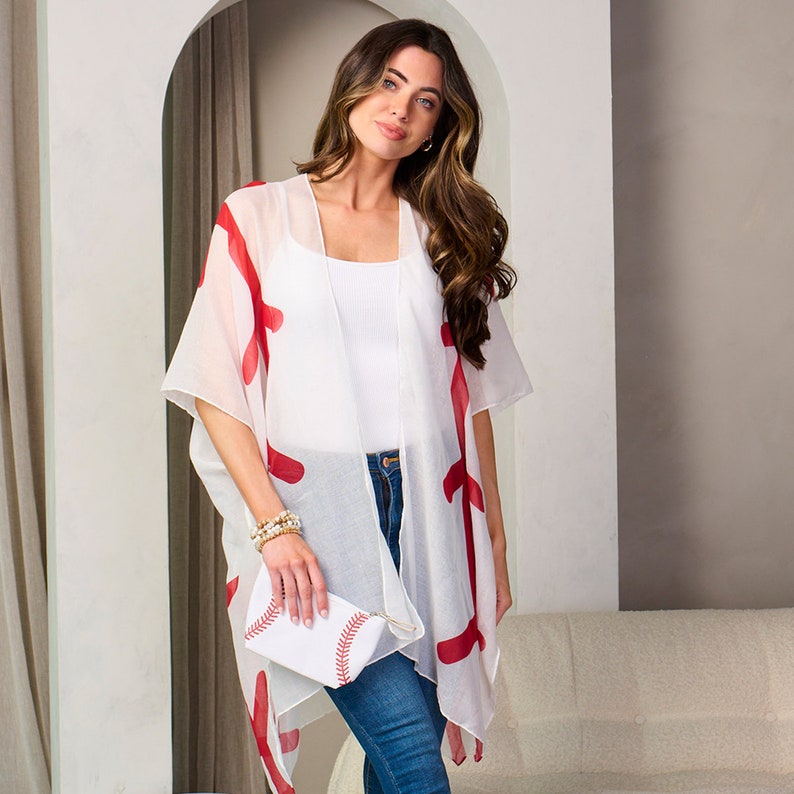 Puede incluir: Un kimono blanco con detalles rojos, combinado con una camiseta blanca y vaqueros azules. Se sostiene un clutch blanco con costuras rojas de b&eacute;isbol. El conjunto es informal y elegante.