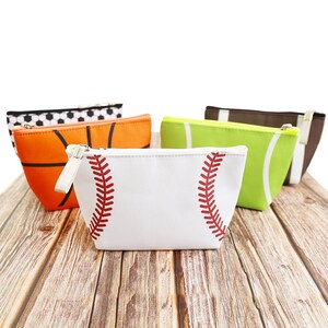 Puede incluir: Una colecci&oacute;n de neceseres con cremallera con dise&ntilde;os deportivos. Las bolsas presentan un b&eacute;isbol, un baloncesto, un bal&oacute;n de f&uacute;tbol, un f&uacute;tbol americano y una pelota de tenis. La bolsa de b&eacute;isbol es blanca con costuras rojas. Las otras bolsas son naranjas, verdes, marrones y negras.