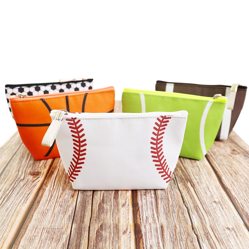 Puede incluir: Una colecci&oacute;n de neceseres con cremallera con dise&ntilde;os deportivos. Las bolsas presentan un b&eacute;isbol, un baloncesto, un bal&oacute;n de f&uacute;tbol, un f&uacute;tbol americano y una pelota de tenis. La bolsa de b&eacute;isbol es blanca con costuras rojas. Las otras bolsas son naranjas, verdes, marrones y negras.