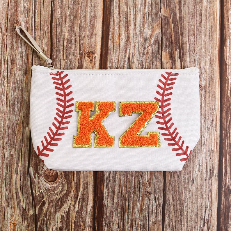 Puede incluir: Bolsa con cremallera blanca con tem&aacute;tica de b&eacute;isbol con detalles de costura rojos. Presenta las letras "KZ" de chenilla naranja y dorada. Incluye una peque&ntilde;a correa para la mu&ntilde;eca. La bolsa est&aacute; sobre un fondo de madera.