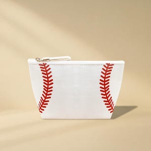 Puede incluir: Bolsa de cosm&eacute;ticos blanca con tem&aacute;tica de b&eacute;isbol con detalles de costuras rojas. La bolsa tiene un cierre de cremallera con una peque&ntilde;a correa blanca y un tirador de cremallera de metal plateado. El dise&ntilde;o imita el aspecto de una pelota de b&eacute;isbol.