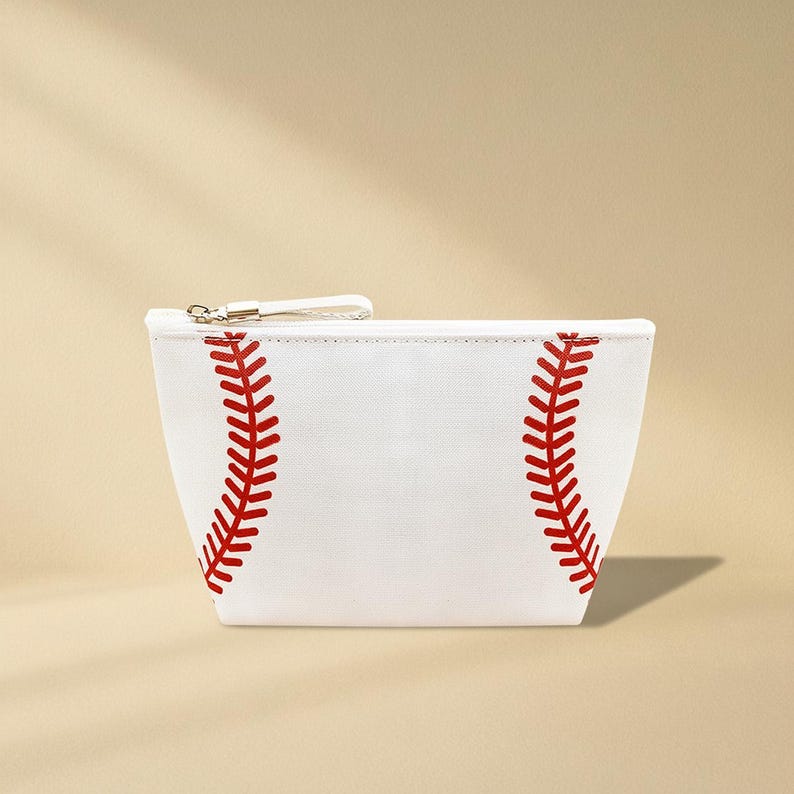 Puede incluir: Bolsa de cosm&eacute;ticos blanca con tem&aacute;tica de b&eacute;isbol con detalles de costuras rojas. La bolsa tiene un cierre de cremallera con una peque&ntilde;a correa blanca y un tirador de cremallera de metal plateado. El dise&ntilde;o imita el aspecto de una pelota de b&eacute;isbol.