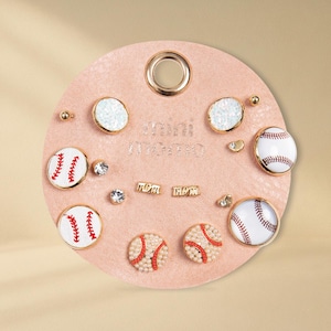 May include: A collection of baseball-themed earrings and studs displayed on a pink circular holder. The earrings feature baseball designs, sparkling accents, and the text "mini momo".