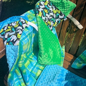 May include: A collection of baby blankets in various shades of blue and green. One blanket features a pattern of video game controllers on a black background. The blankets are made of soft, textured fabric and are folded on a wooden bench.