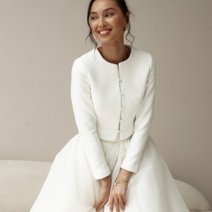 May include: A white bridal gown with a fitted bodice and a full skirt. The gown features a long-sleeved jacket with pearl buttons.